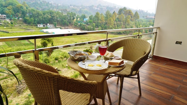 A balcony with wicker chairs, a dining table with food and drinks, and a view of a green landscape with buildings in the distance. - Nakshville  (Bhimtal)