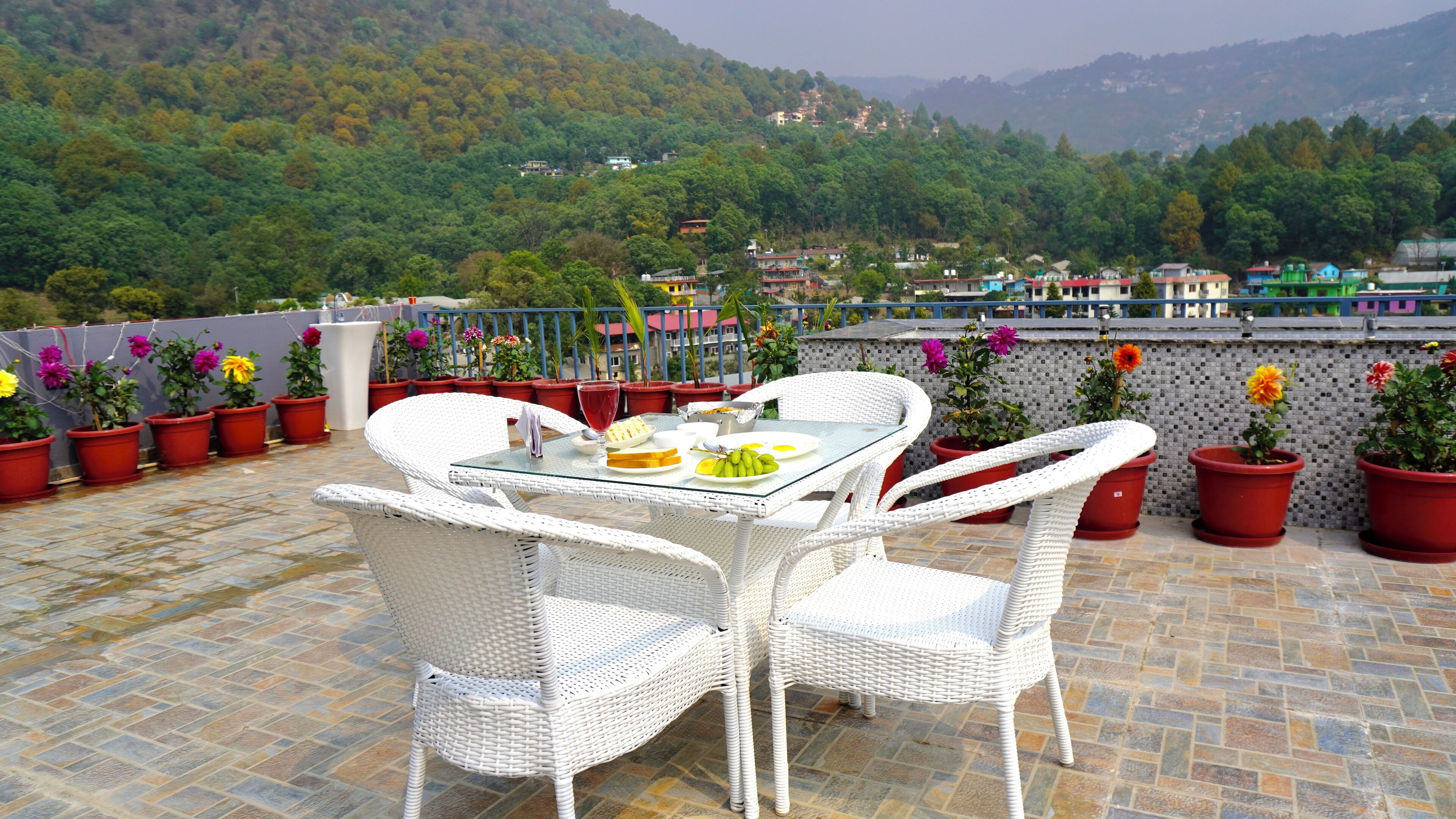 A terrace with a table and chairs, surrounded by potted plants and a scenic view of a village and mountains.