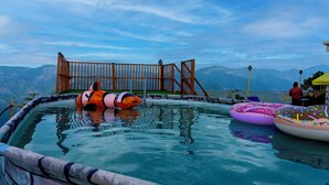 A swimming pool with inflatable toys and a wooden deck overlooking a mountainous landscape. - Orchard Green Shimla (Shimla)