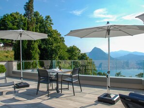 A terrace with two tables, chairs, and umbrellas, overlooking a scenic landscape with mountains and a body of water.