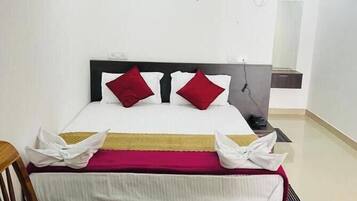 A hotel room with a bed, a desk with a kettle and breakfast tray, a chair, and an air conditioning unit.