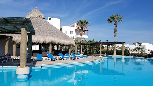 A swimming pool with lounge chairs, a thatched-roof structure, and palm trees. - Contemporary Conveniently Located 1 BR condo, Walk to beach, restaurants & bars. (San José del Cabo)