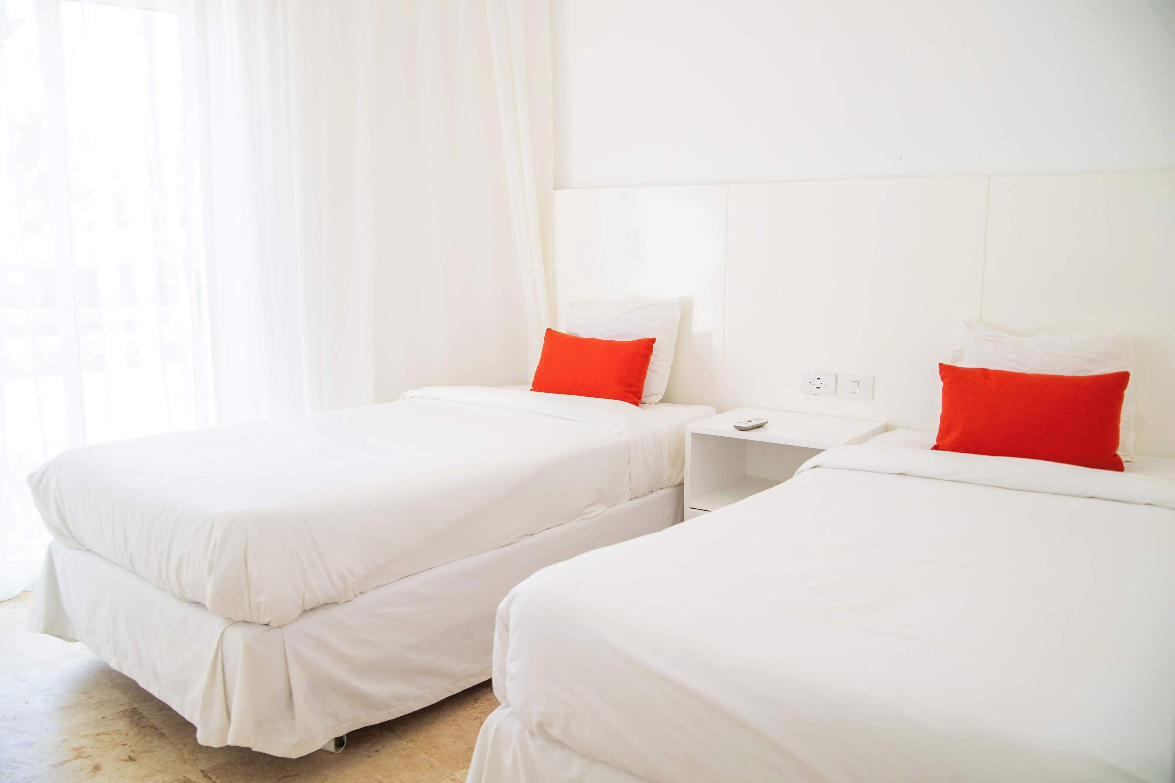 A hotel room with two beds, each with white bedding and red pillows, and a white nightstand with a remote control.
