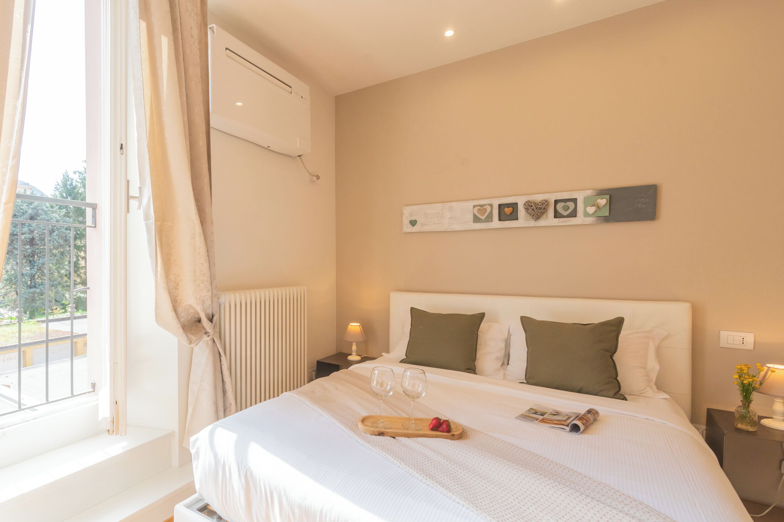 A neatly made bed with a tray of two glasses and strawberries, a bedside table with a lamp, and a wall-mounted decoration.