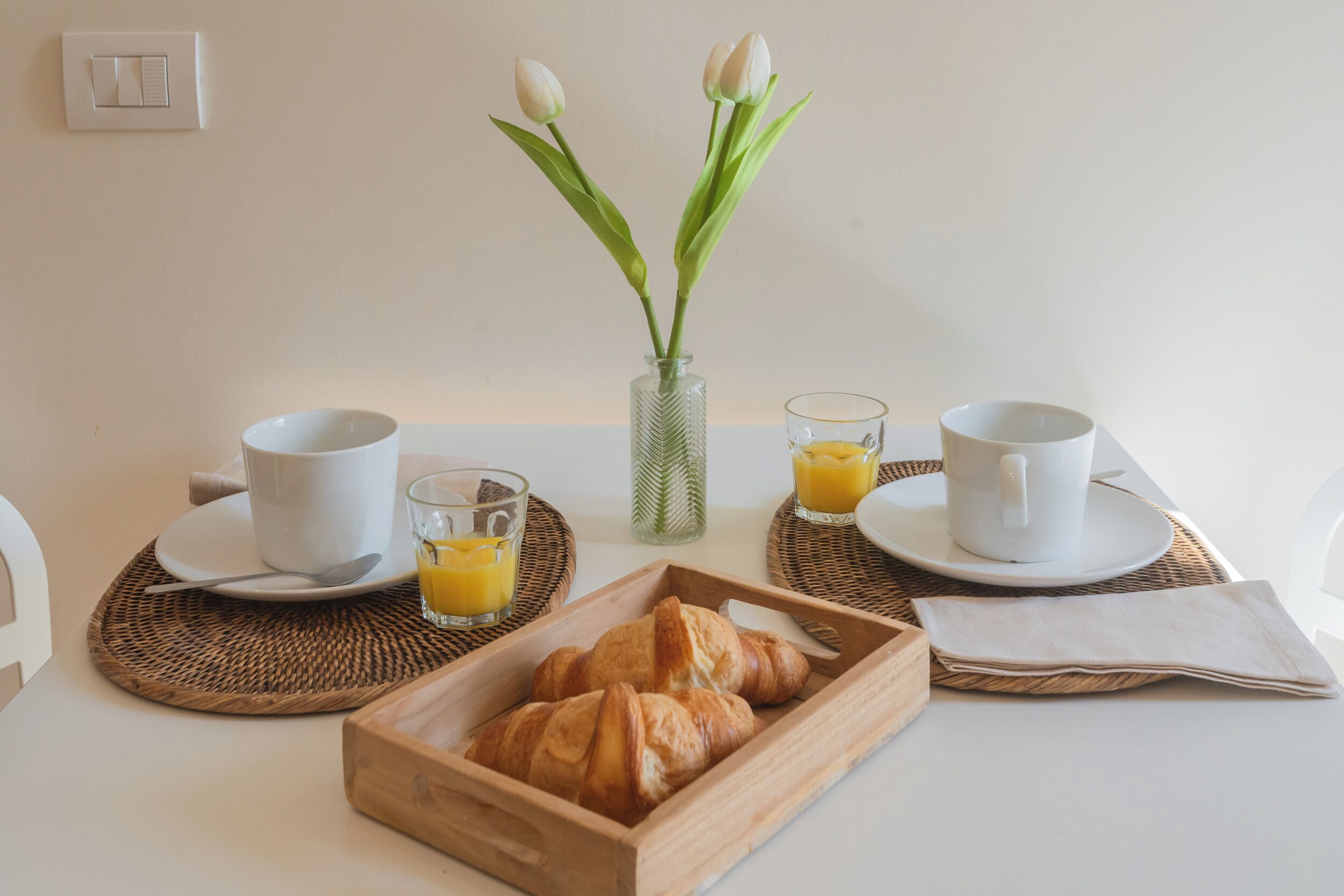 Une table mise pour le petit-déjeuner avec des croissants, deux tasses blanches, deux verres de jus d’orange et un vase contenant des tulipes blanches.