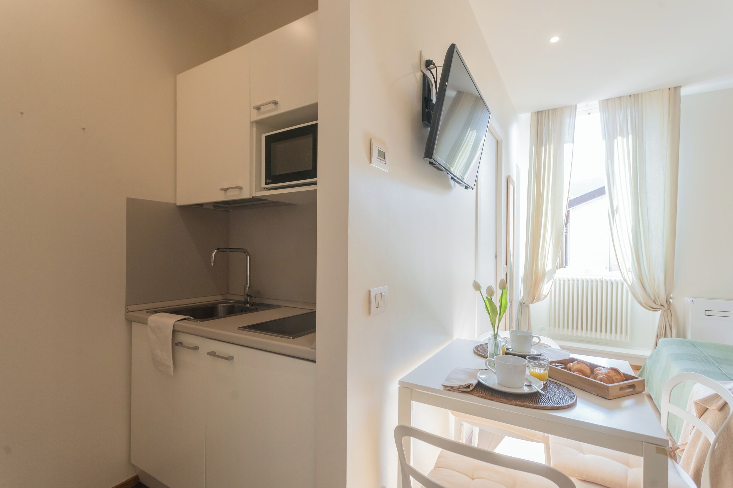 A compact kitchenette with a microwave, sink, and cabinets. A small dining table with a chair is positioned near a window with curtains.