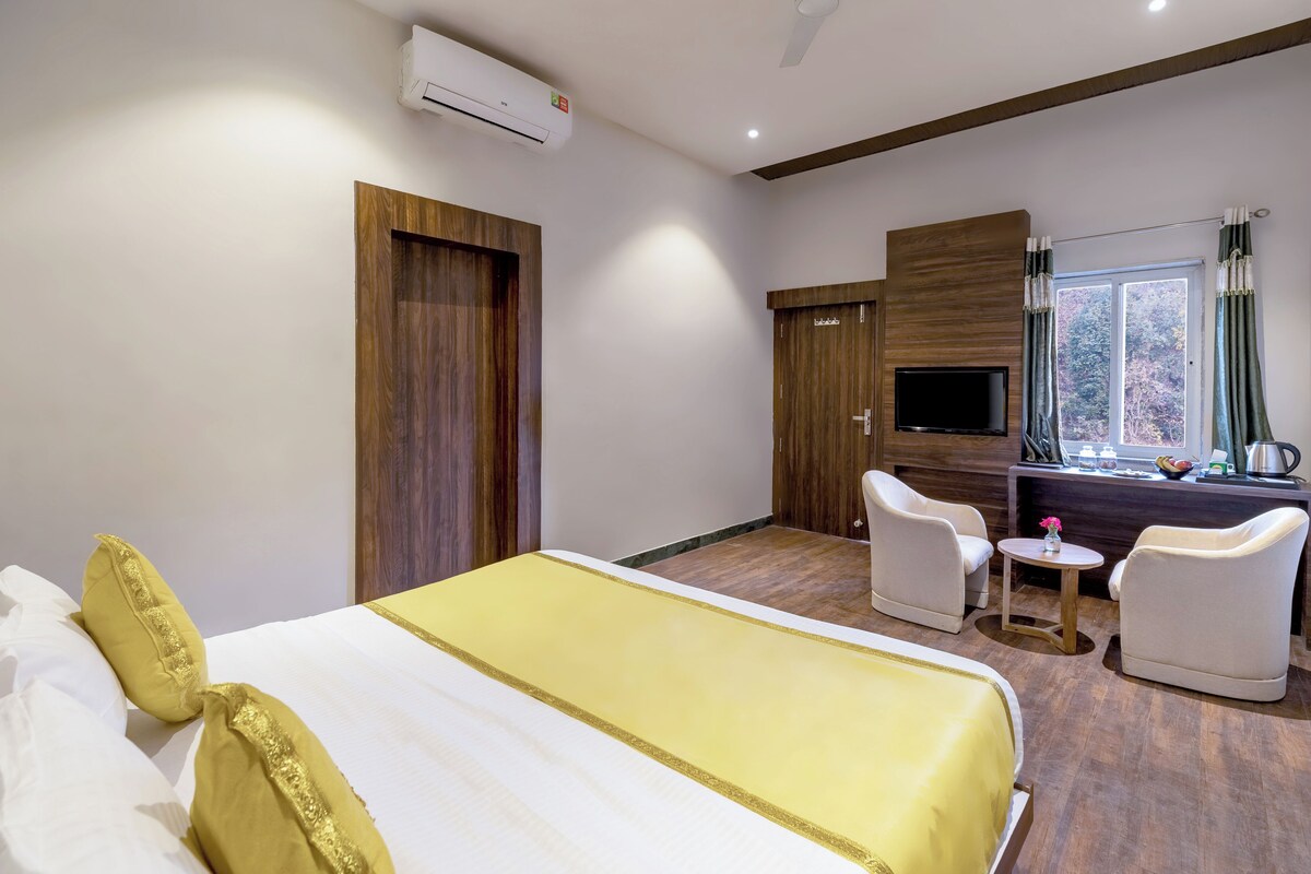 A hotel room with a bed, two white armchairs, a small table, a TV, and a window with curtains.