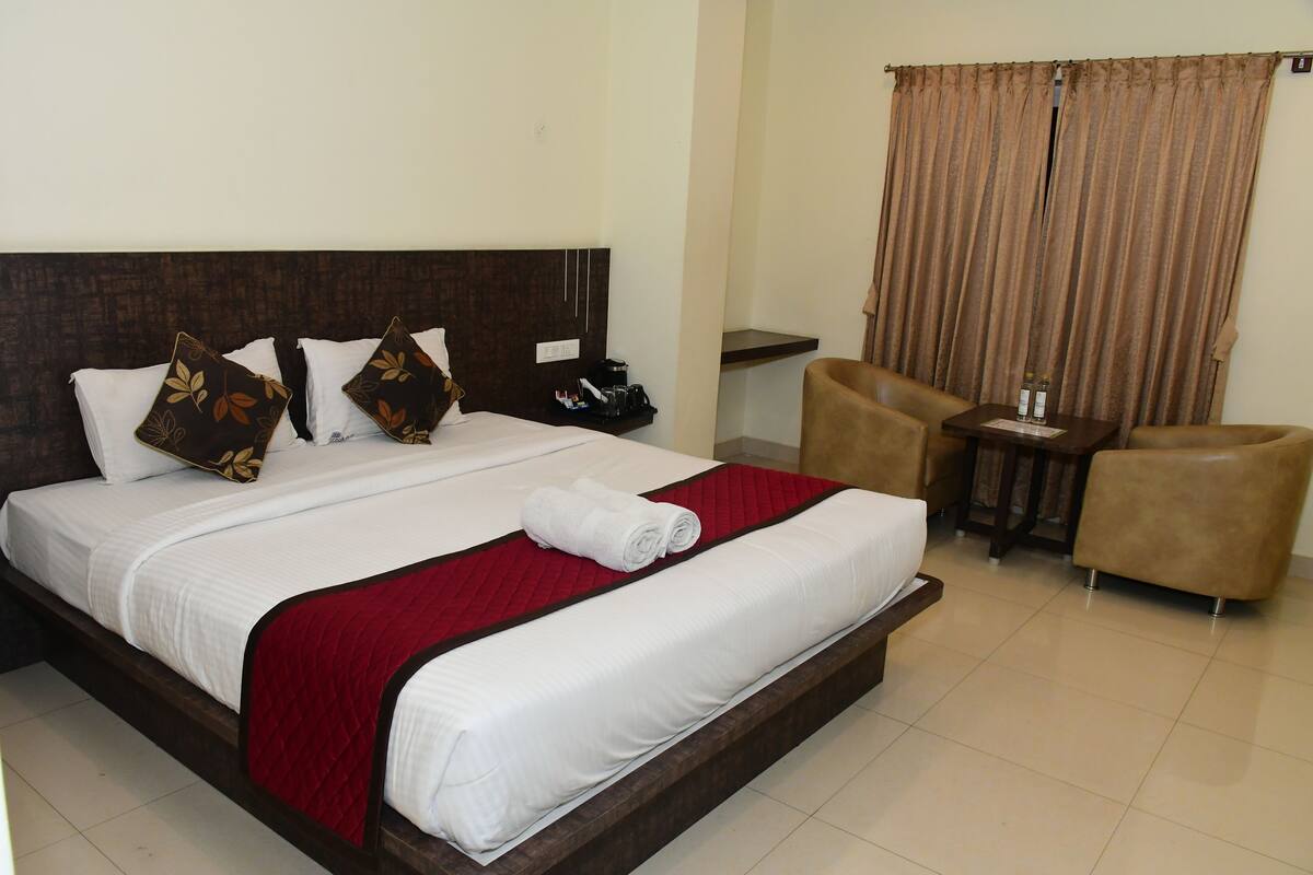 Quarto de hotel com uma cama grande, um cobertor vermelho, duas almofadas marrons, uma mesinha com garrafas e duas cadeiras.