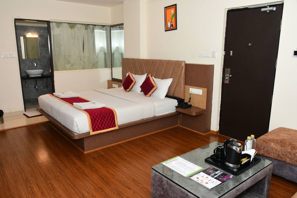 Quarto de hotel com cama, mesa de centro de vidro, porta e banheiro visível através de um painel de vidro.