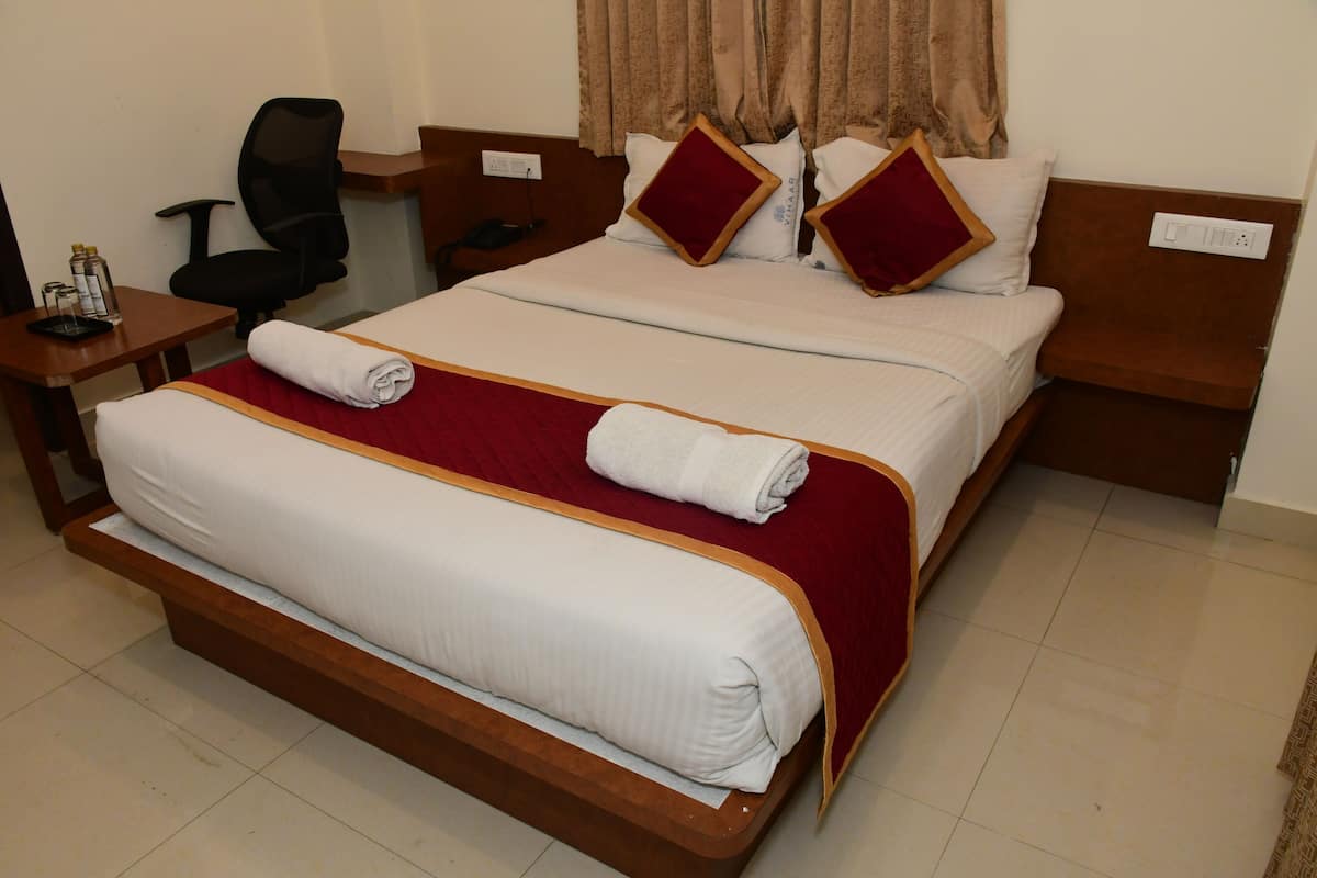 Uma cama de casal bem arrumada com colcha vermelha e roupa de cama branca, uma mesinha de cabeceira de madeira com abajur e uma cadeira de escritório preta.