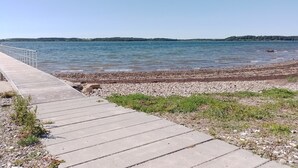 Un chemin de béton mène à une plage de galets, d’où l’on a une vue dégagée sur l’eau et sur la côte au loin.