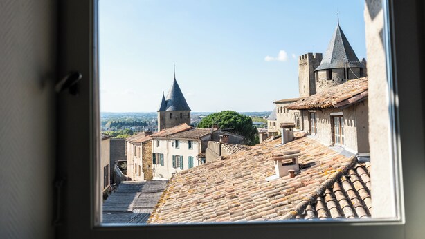 A view through a window of a building with traditional architecture, including stone buildings with tiled roofs and a prominent tower. - Hostel Coeur de Cité (Carcassonne)