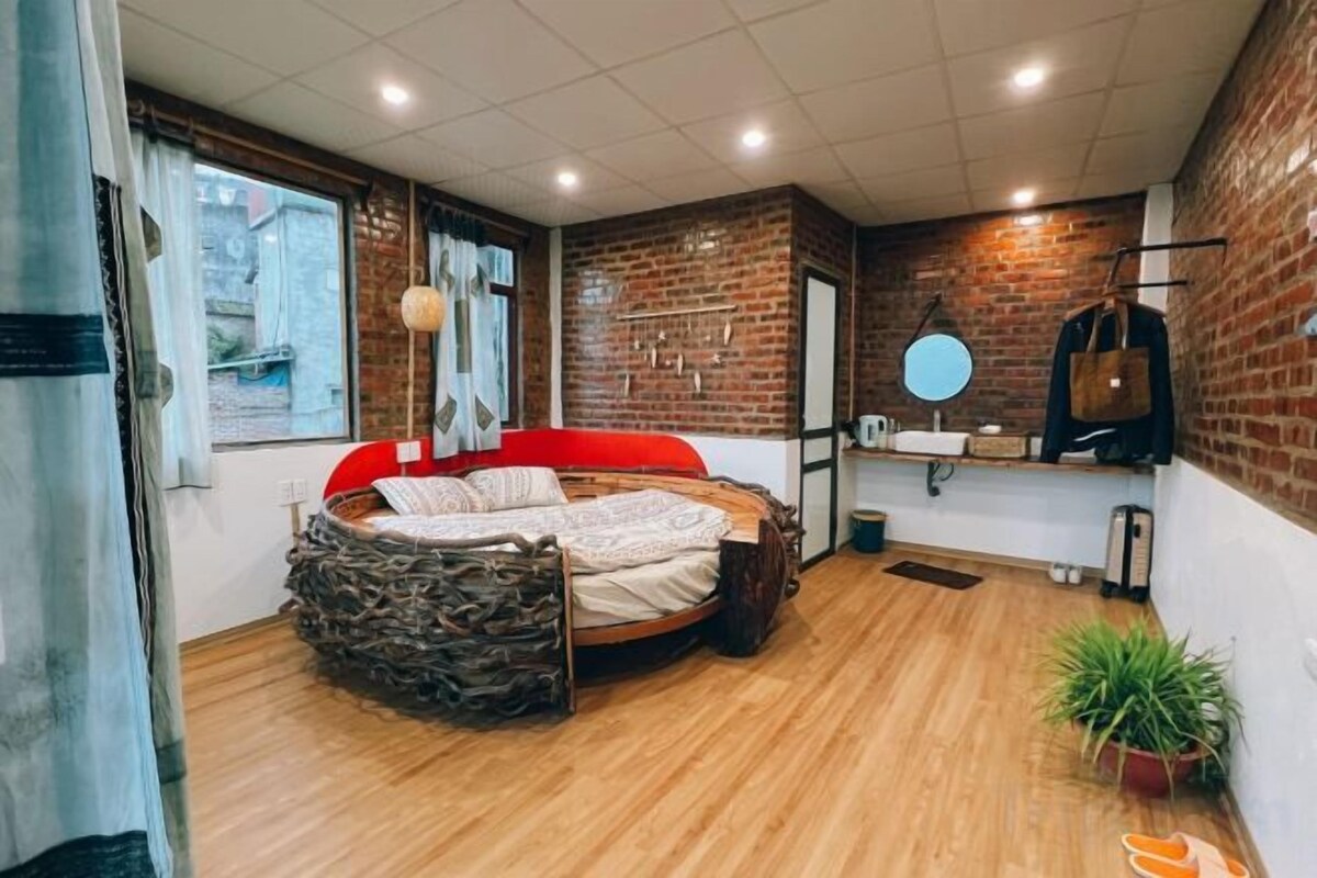 A room with a circular stone bed, a brick wall, a wooden floor, a window with curtains, and a small potted plant.