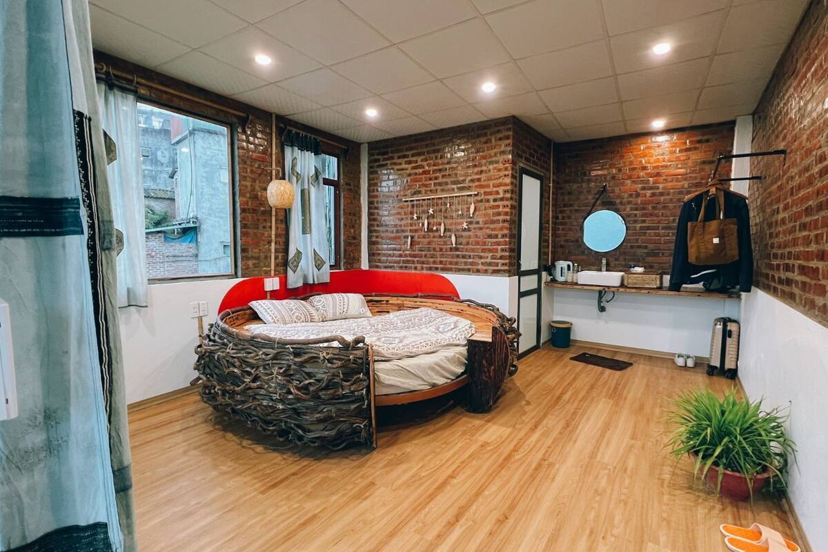 A bedroom with a stone headboard, a red bed, a brick wall, a wooden floor, a window with a view, and a small plant.