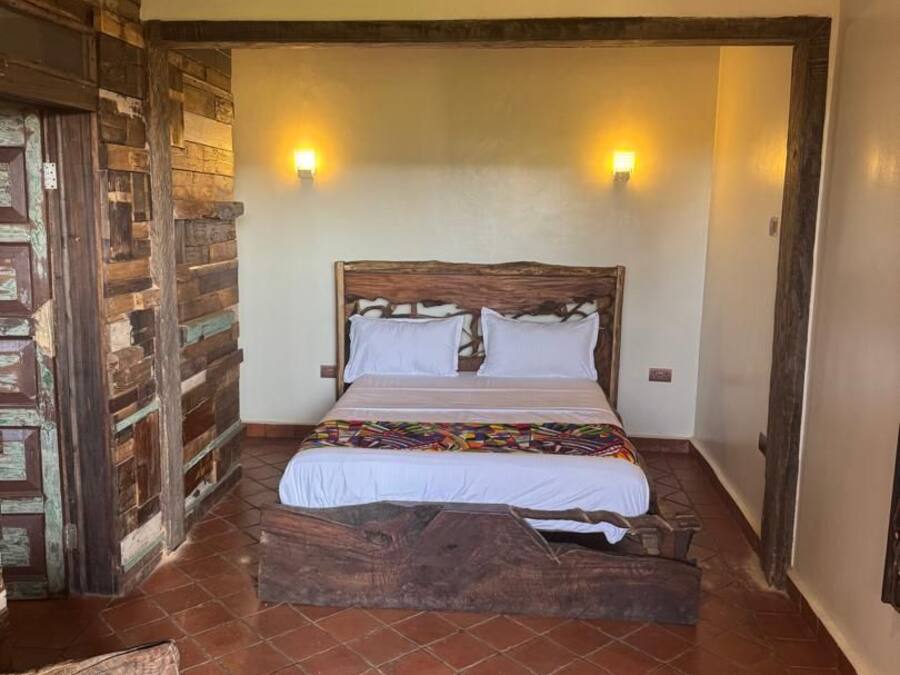 A bedroom with a wooden headboard, a bed with white linens, two wall-mounted lights, and a rustic brick wall.