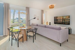 Una sala de estar moderna con una mesa redonda de madera, sillas negras, un sofá lila claro, un televisor de pantalla plana grande y vistas al exterior a través de amplias ventanas.
