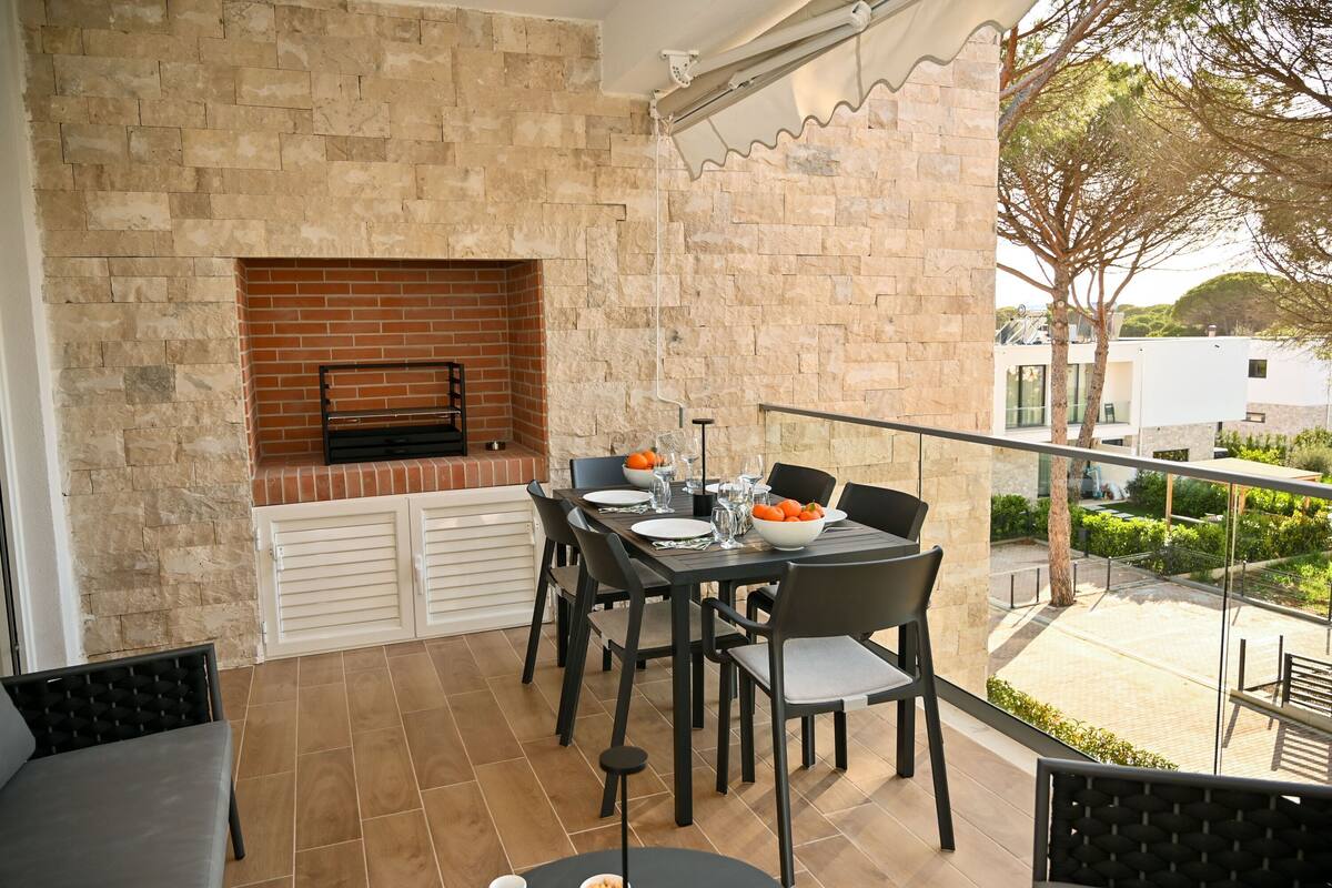 Varanda com mesa de jantar posta para quatro pessoas, lareira de tijolos e vista para um jardim e edifícios.