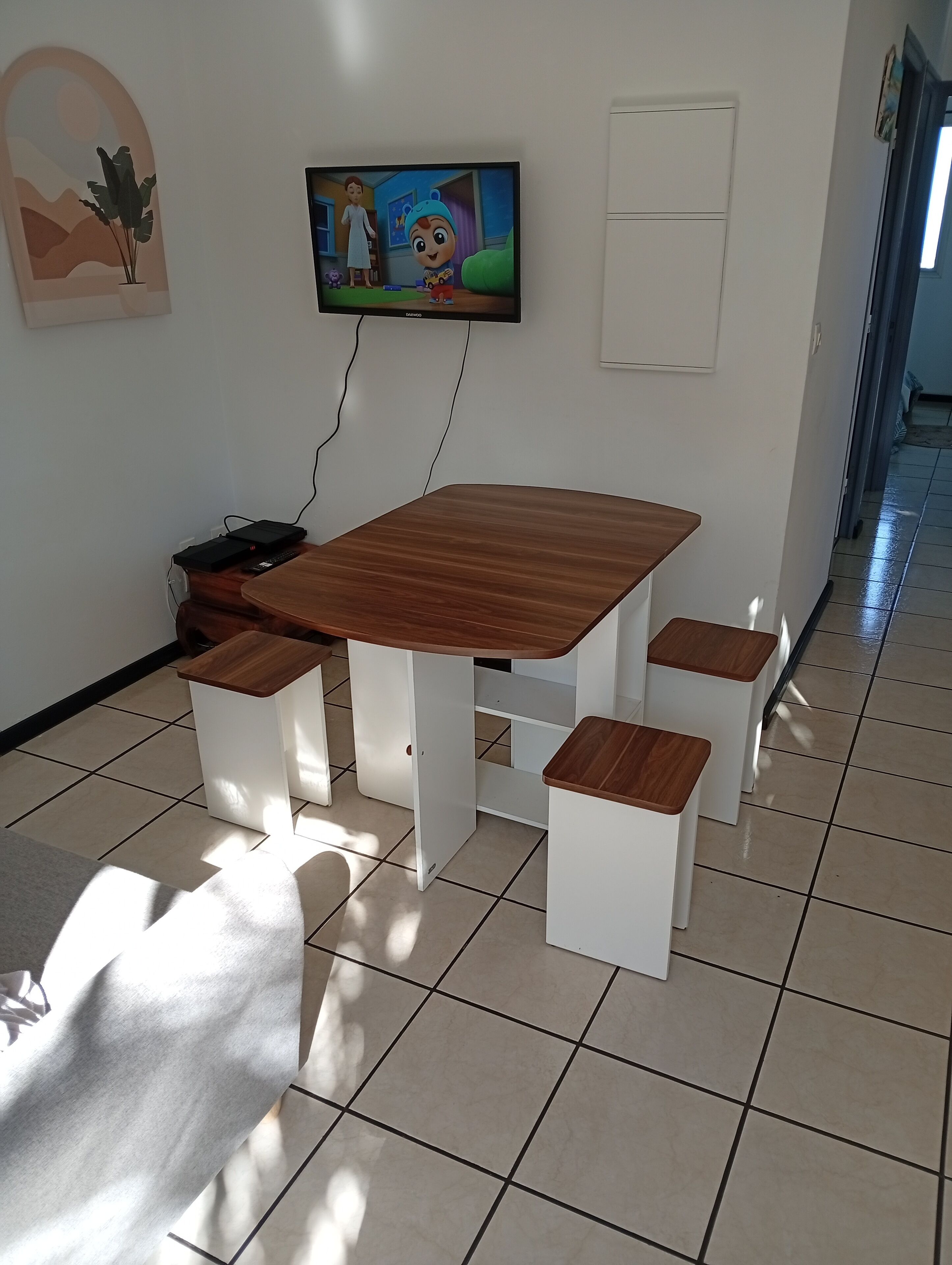 Un comedor con una mesa de madera y bancos, un televisor montado en la pared y una planta en maceta.
