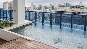 Piscina infinita en la azotea con vistas a la ciudad, rodeada por una terraza de madera y barandas de vidrio.