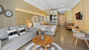 A modern hotel room with a sofa, a round wooden table, a bed, a TV, and a dining area. - Rivana by Kozystay - Nusa Dua (Nusa Dua)