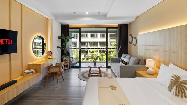 A modern hotel room with a large bed, a sofa, a TV, and a balcony with a view of buildings. - Rivana by Kozystay - Nusa Dua (Nusa Dua)