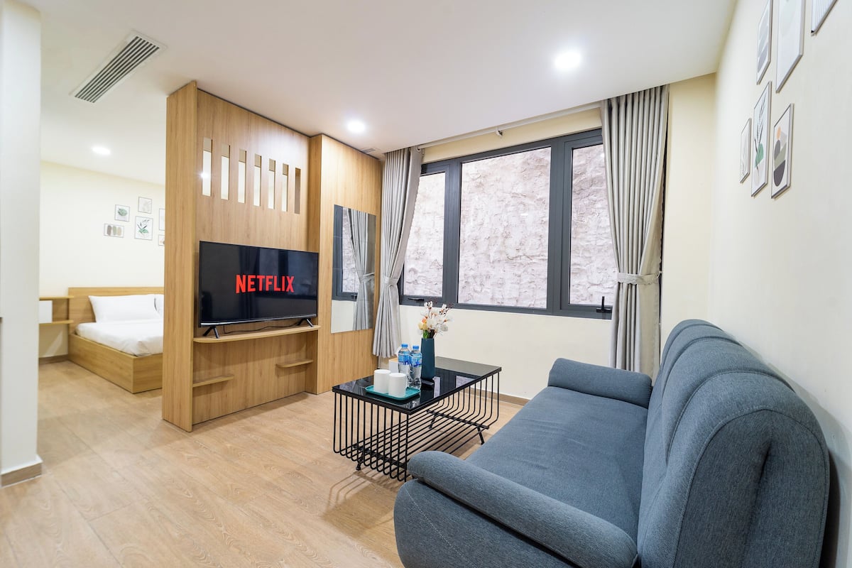 Une chambre d’hôtel moderne avec un lit en bois, un canapé gris, un téléviseur à écran plat et une petite table sur laquelle est posé un vase de fleurs.
