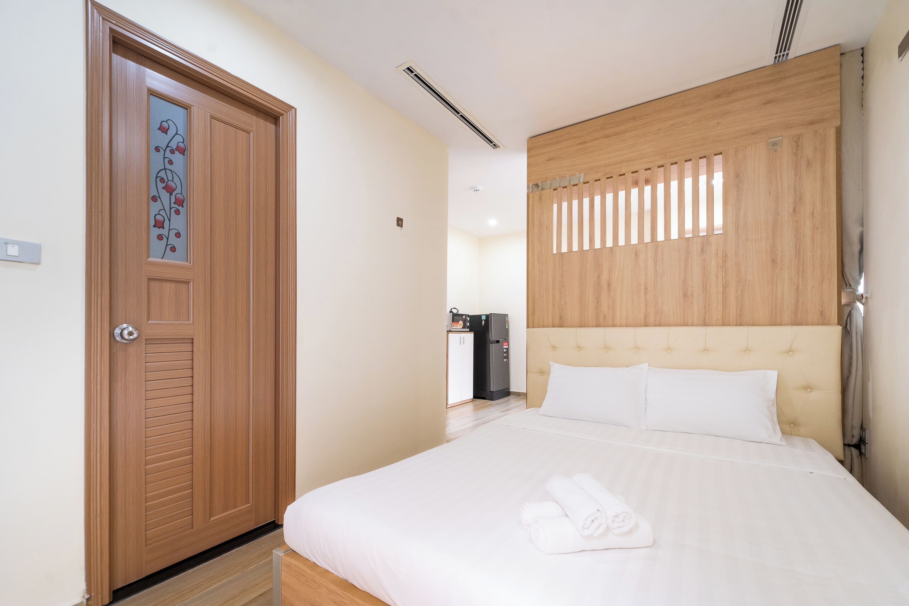 Ein Hotelzimmer mit Holztür, einem Bett mit weißer Bettwäsche, einem Kopfteil mit vertikalen Lamellen und einer kleinen Küchenzeile im Hintergrund.