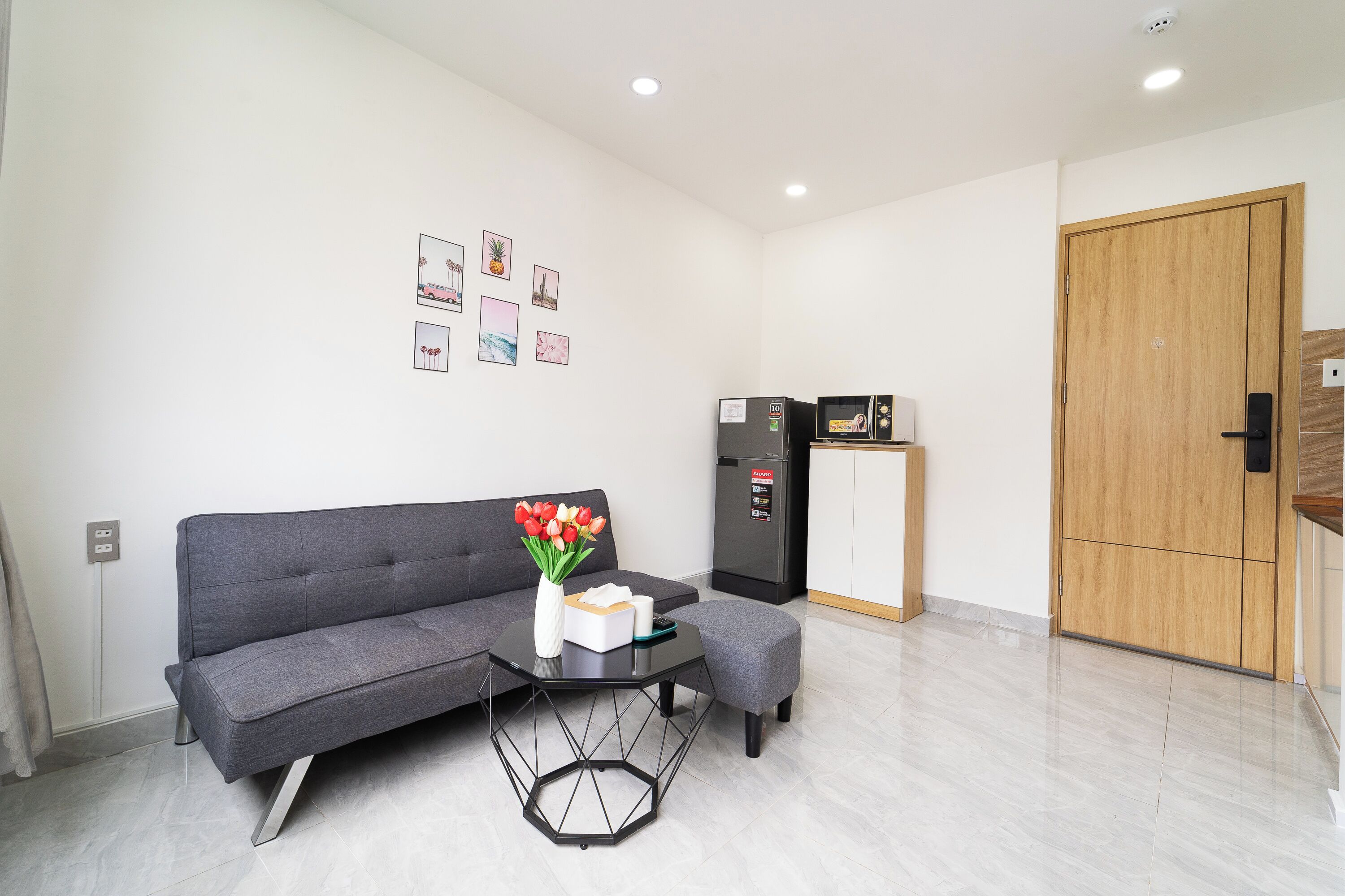 Een moderne woonkamer met een grijze bank, een zwarte salontafel met een vaas rode tulpen, en een klein kastje met een koelkast.