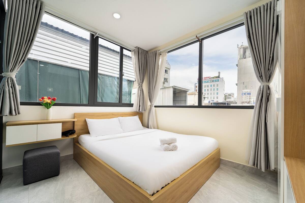 Une chambre d’hôtel moderne avec un grand lit, une table de chevet, un petit tabouret et une fenêtre avec des rideaux.