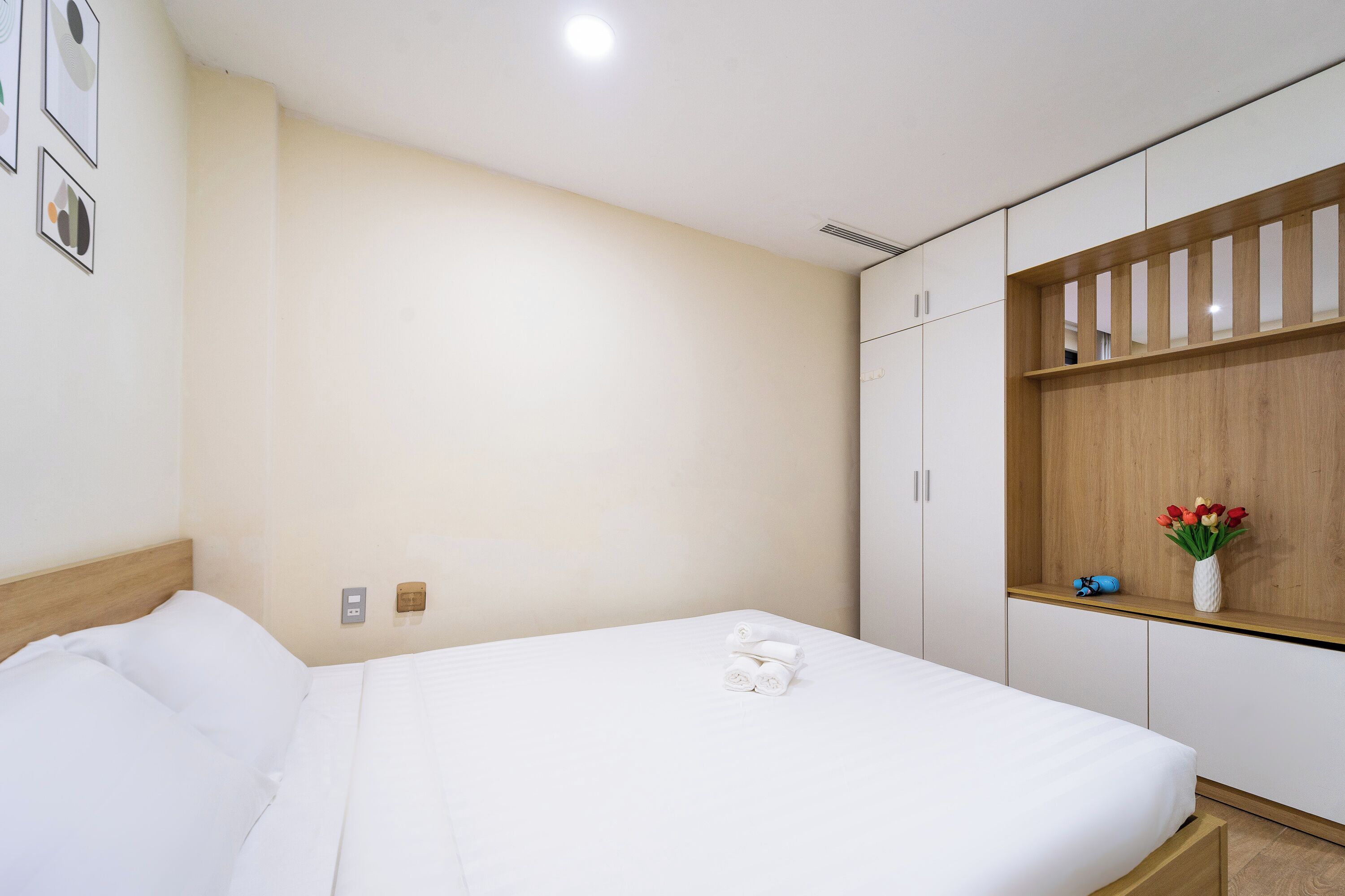 A neatly made bed with white linens, a wooden headboard, and a small shelf with a vase of flowers.