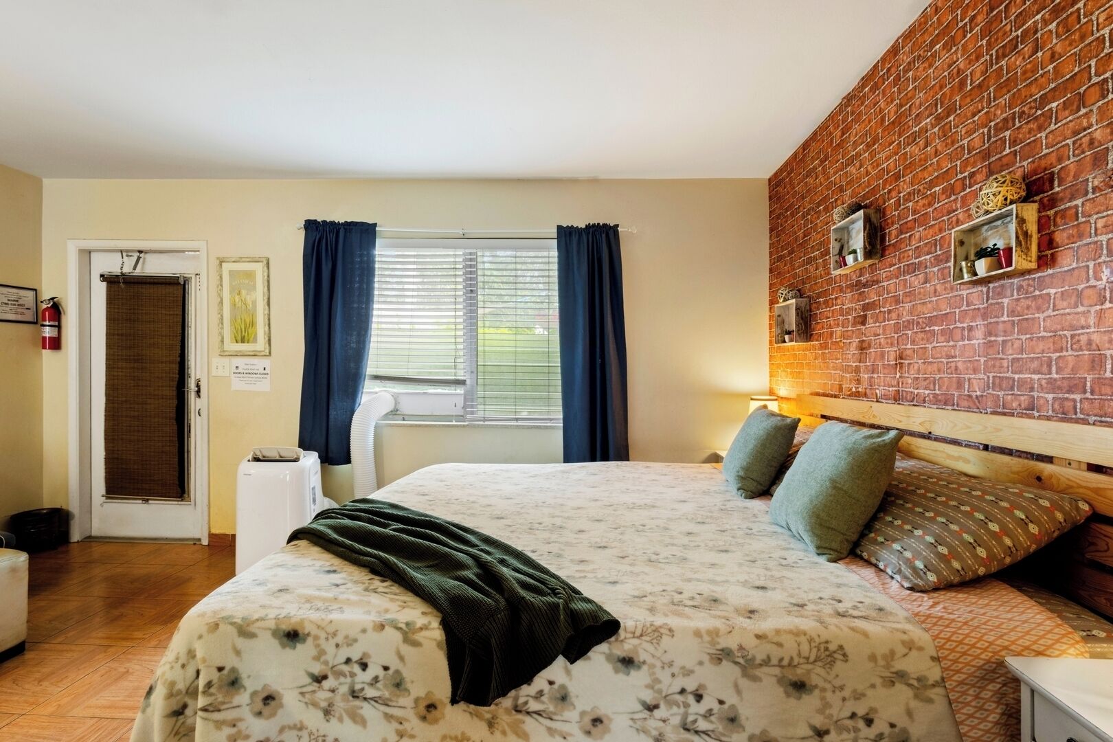 A bedroom with a bed, a brick wall, a window with curtains, and a door.
