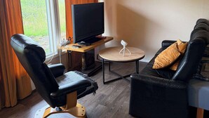 A room with a TV on a wooden stand, a black leather sofa, a round wooden coffee table, and a wooden floor.