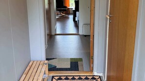A hallway with a wooden bench, a welcome mat with a bear design, and a view into a room with a TV and a window with curtains.