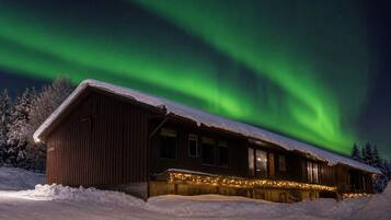 Une cabane en bois, dont le toit est recouvert de neige, entourée de neige, sous un ciel nocturne illuminé par des aurores boréales.
