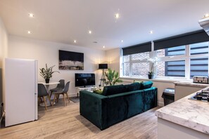 A modern living room with a dark green sofa, a dining area with a table and chairs, a flat-screen TV, and a kitchen with a stove. - Haymarket House (Basingstoke)