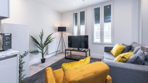 A modern living room with a grey sofa, yellow cushions, a black TV on a wooden stand, a floor lamp, and a potted plant. - Haymarket House (Basingstoke)