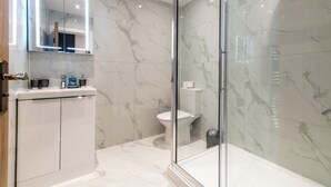 A modern bathroom with a glass-enclosed shower, a white toilet, and a vanity with a mirror and lights. - Haymarket House (Basingstoke)