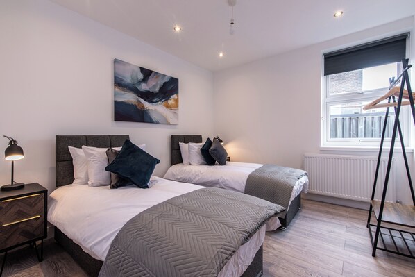 A modern bedroom with two beds, a painting on the wall, a radiator, and a window with a roller blind. - Haymarket House (Basingstoke)