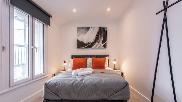 A modern bedroom with a large bed, two bedside tables with lamps, a window with blinds, and a painting on the wall. - Haymarket House (Basingstoke)