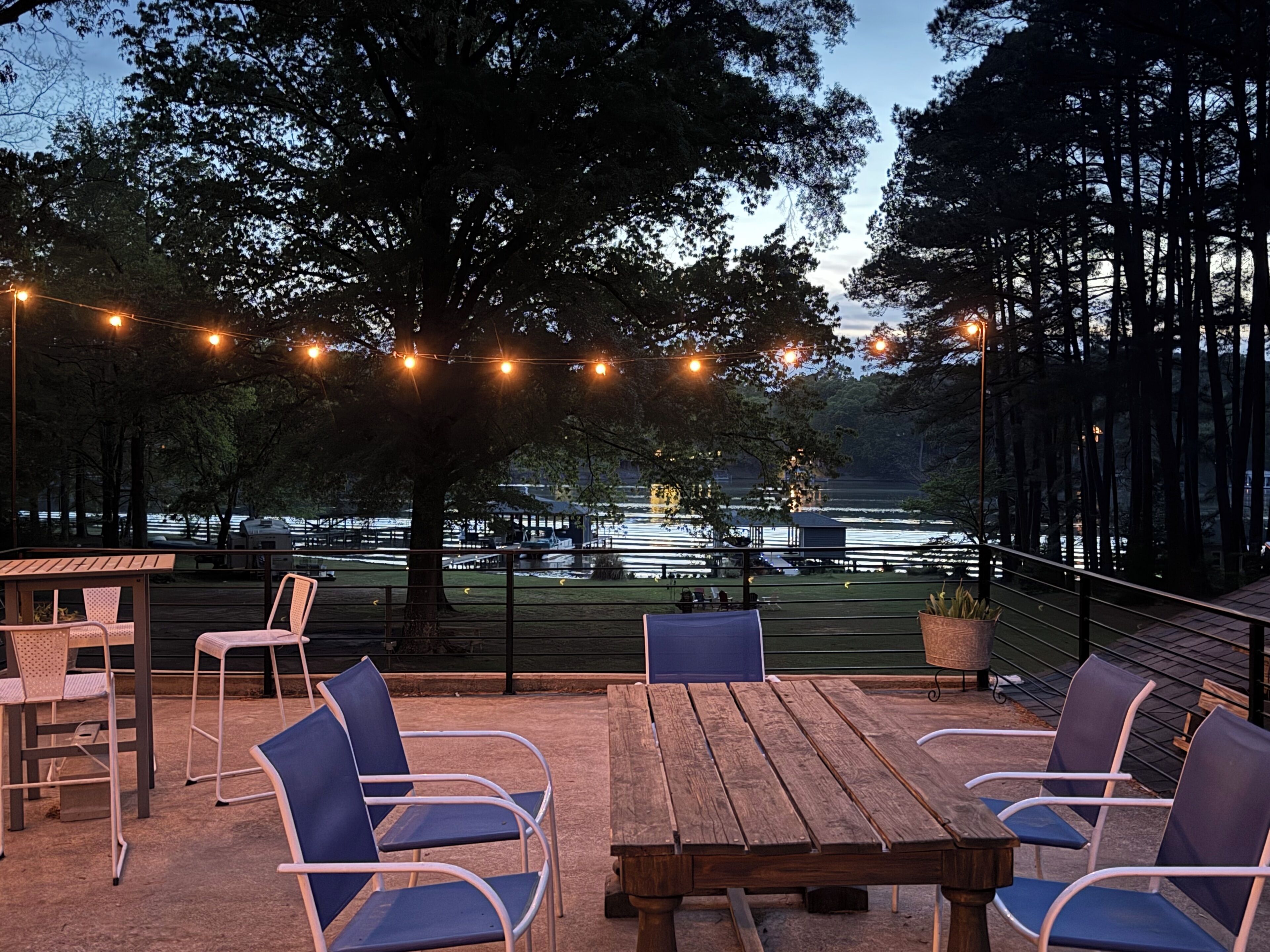 An outdoor patio with wooden tables and chairs, string lights, and a view of a lake with boats.