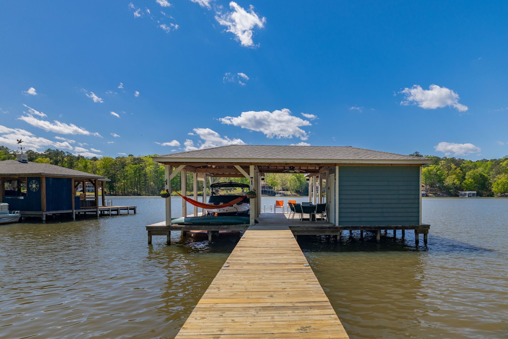 A wooden dock leading to a covered seating area with a hammock and tables, surrounded by calm water and trees.
