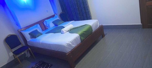 Un lit simple avec un cadre en bois, une chaise et un mur bleu orné d’un luminaire.