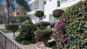 Un bâtiment moderne à l’extérieur blanc, doté d’un balcon avec des garde-corps en verre et d’un jardin bien entretenu, agrémenté de divers arbustes et d’un palmier.