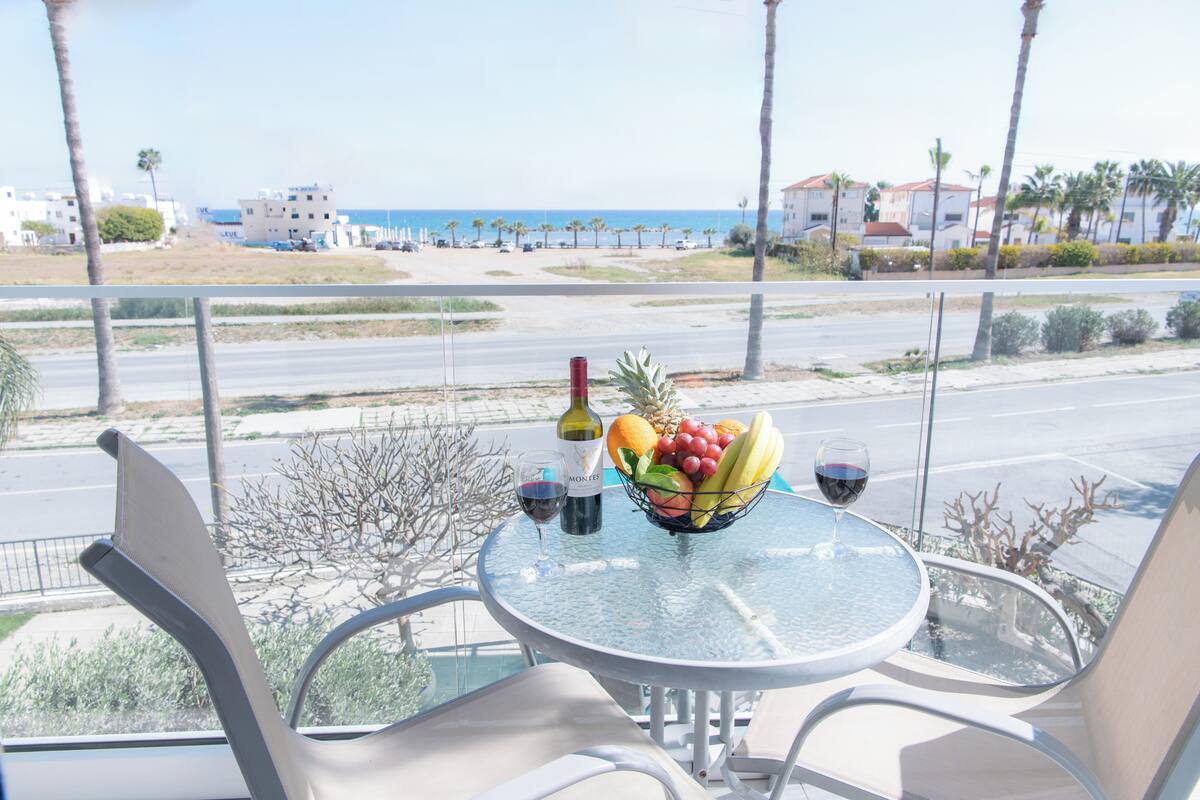 Un balcón con una mesa dispuesta para dos personas, que incluye un frutero, copas de vino y una botella. La vista muestra una playa y palmeras.