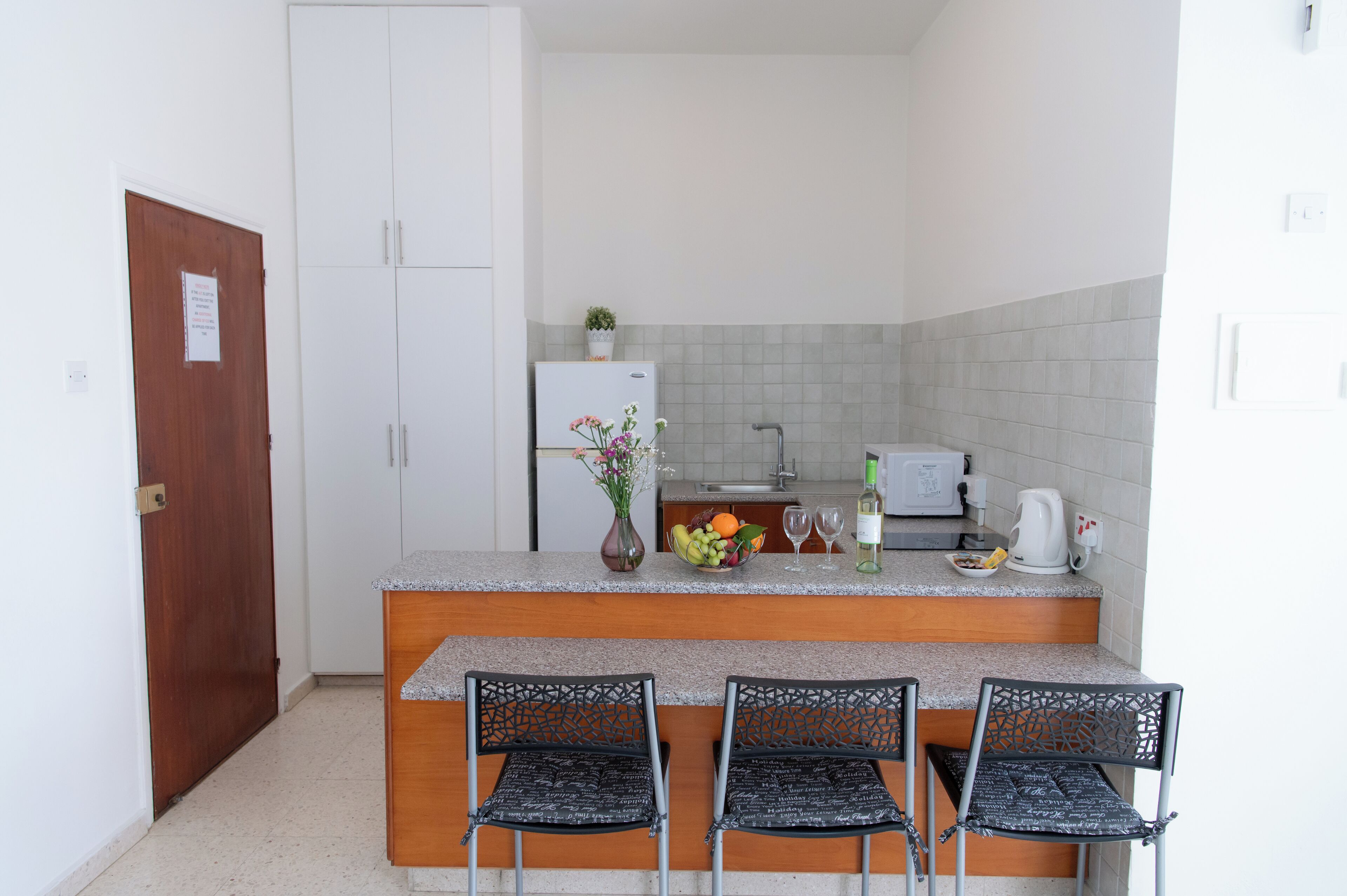 Une petite cuisine équipée d’un réfrigérateur, d’un four à micro-ondes et d’un évier. On y trouve quatre chaises et une petite table sur laquelle est posé un vase de fleurs.