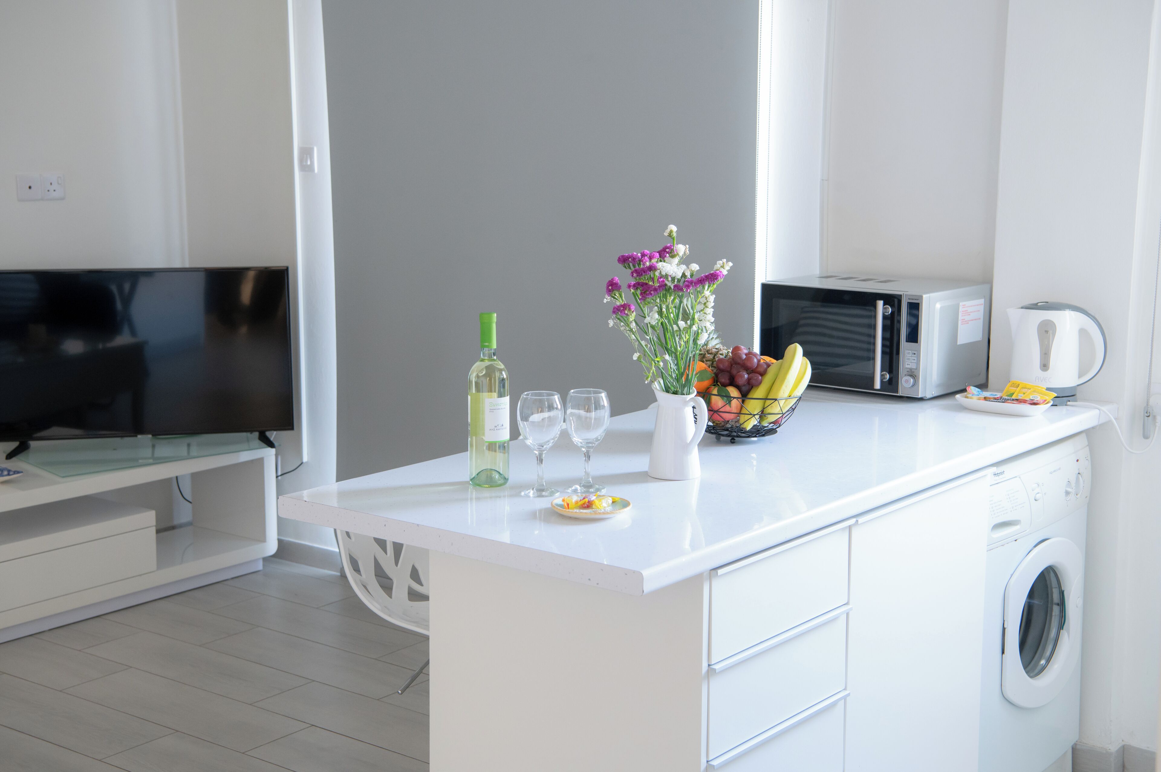 A modern kitchen with a white countertop, a washing machine, a microwave, a television, a bottle of wine, two glasses, and a fruit bowl.