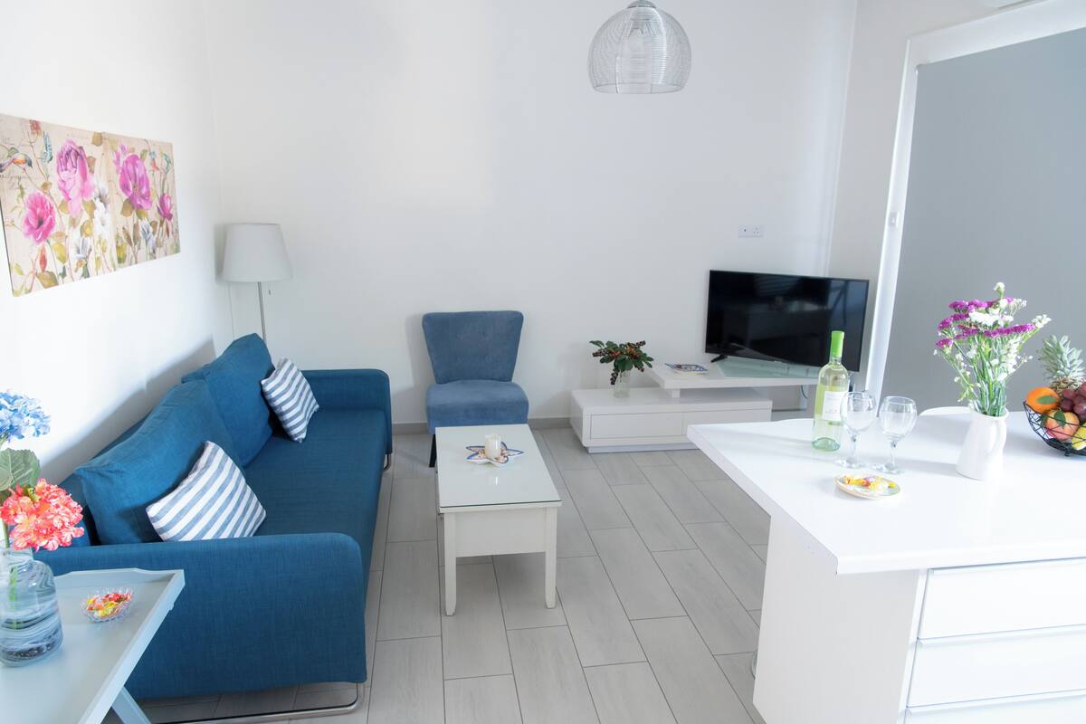 Una sala moderna con un sofá azul, una mesita blanca pequeña, un sillón azul, un televisor y una zona de cocina con encimera blanca y varios objetos.