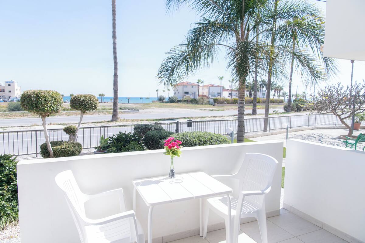 Un balcón con sillas y una mesa blancos, con vista a una playa y palmeras.
