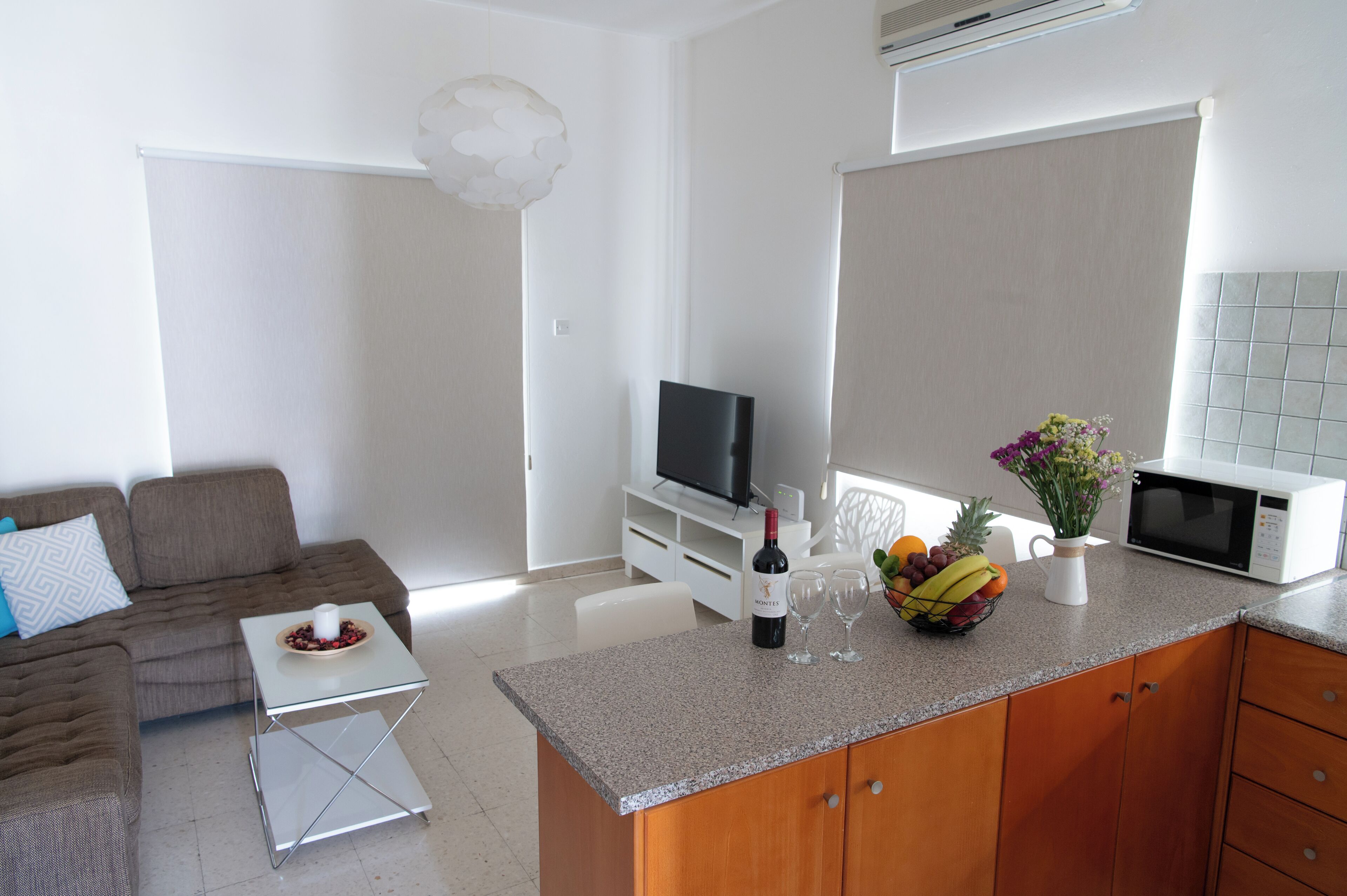A modern living room with a sofa, coffee table, and a television. A kitchen area with a microwave and a fruit bowl is visible.
