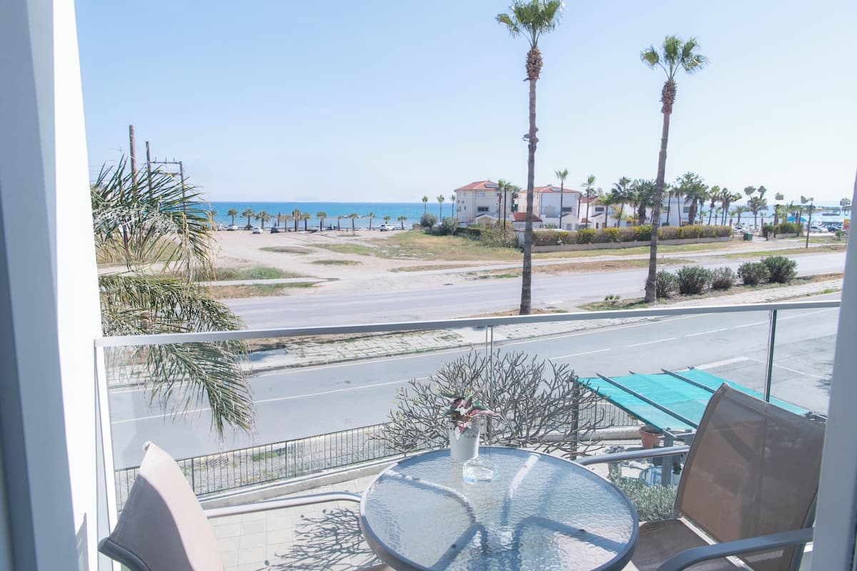 Un balcón con mesa y sillas, con vistas a una playa con palmeras y un cielo despejado.